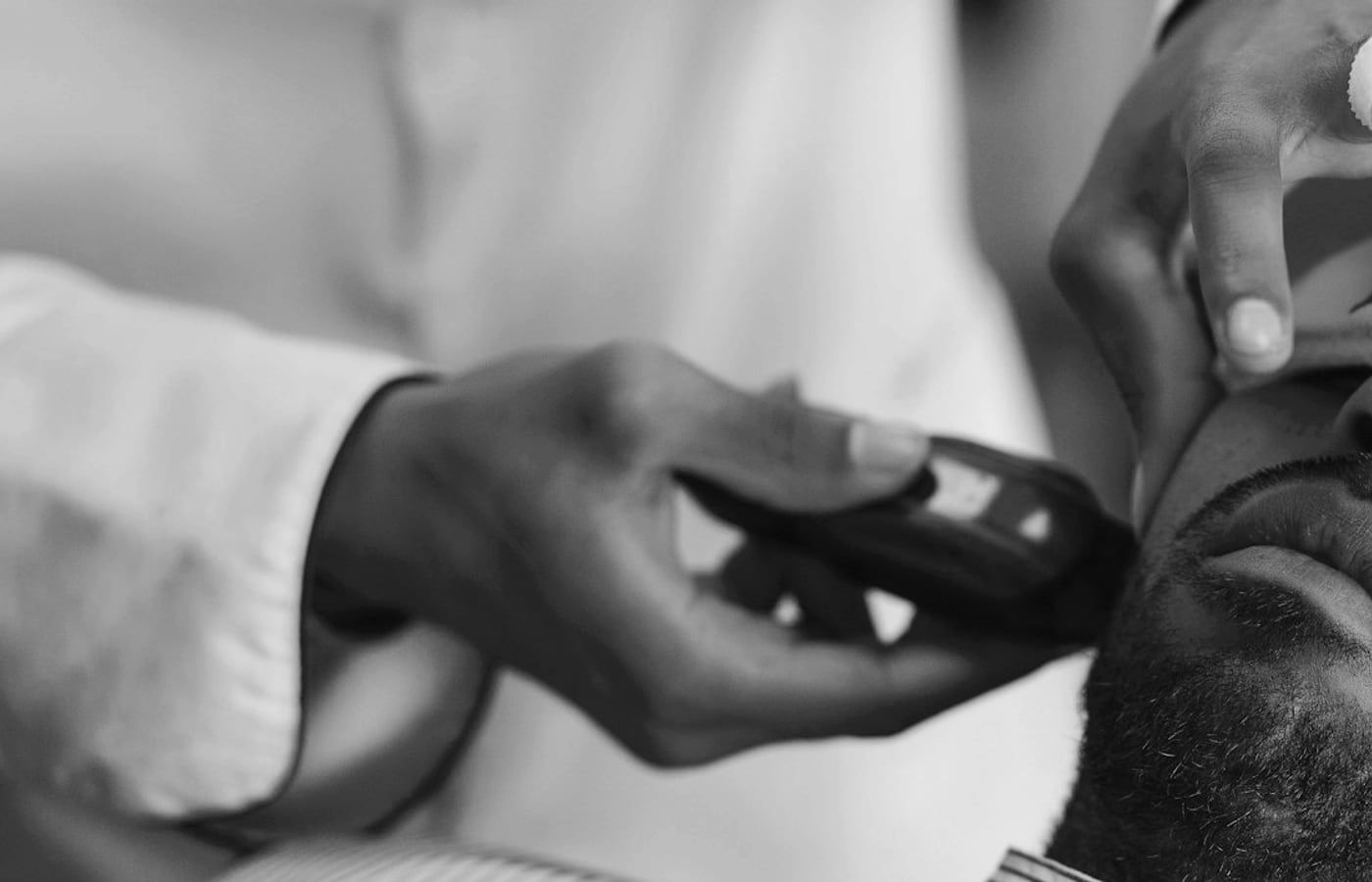 A barber's hands holding a trimmer against a client's jaw, sleeve rolled, mid-cut.