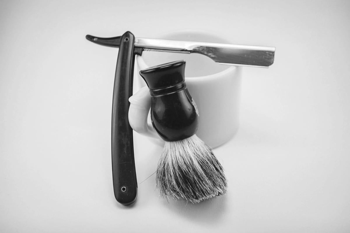 An open razor resting across a soap mug beside a badger shaving brush.